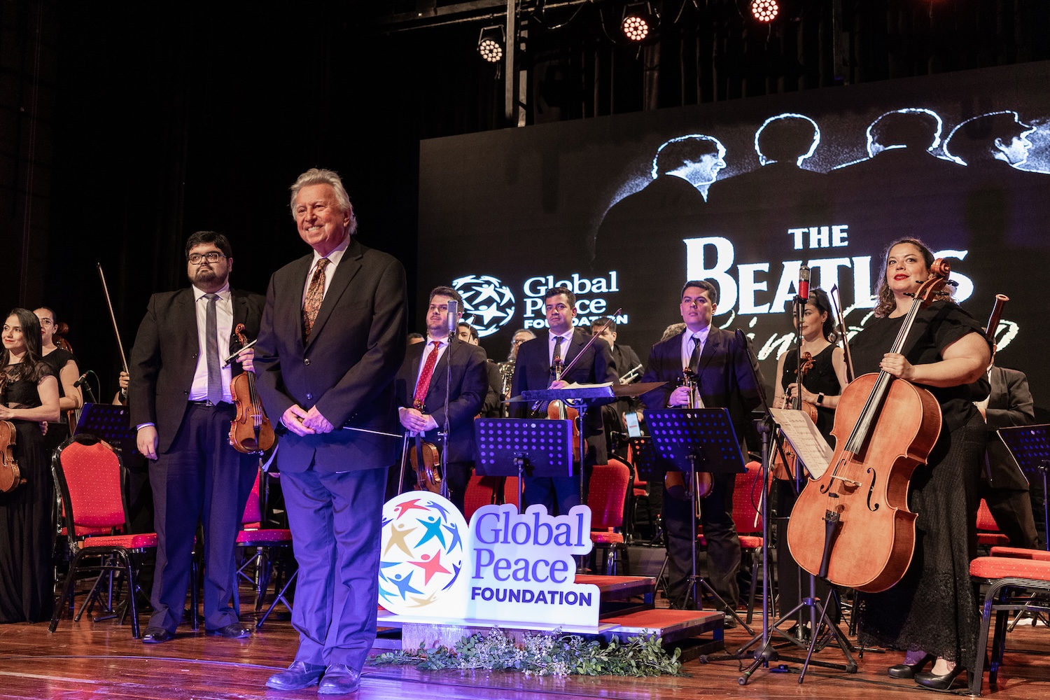An orchestra graces the stage, led by a conductor, with musicians poised to play. A