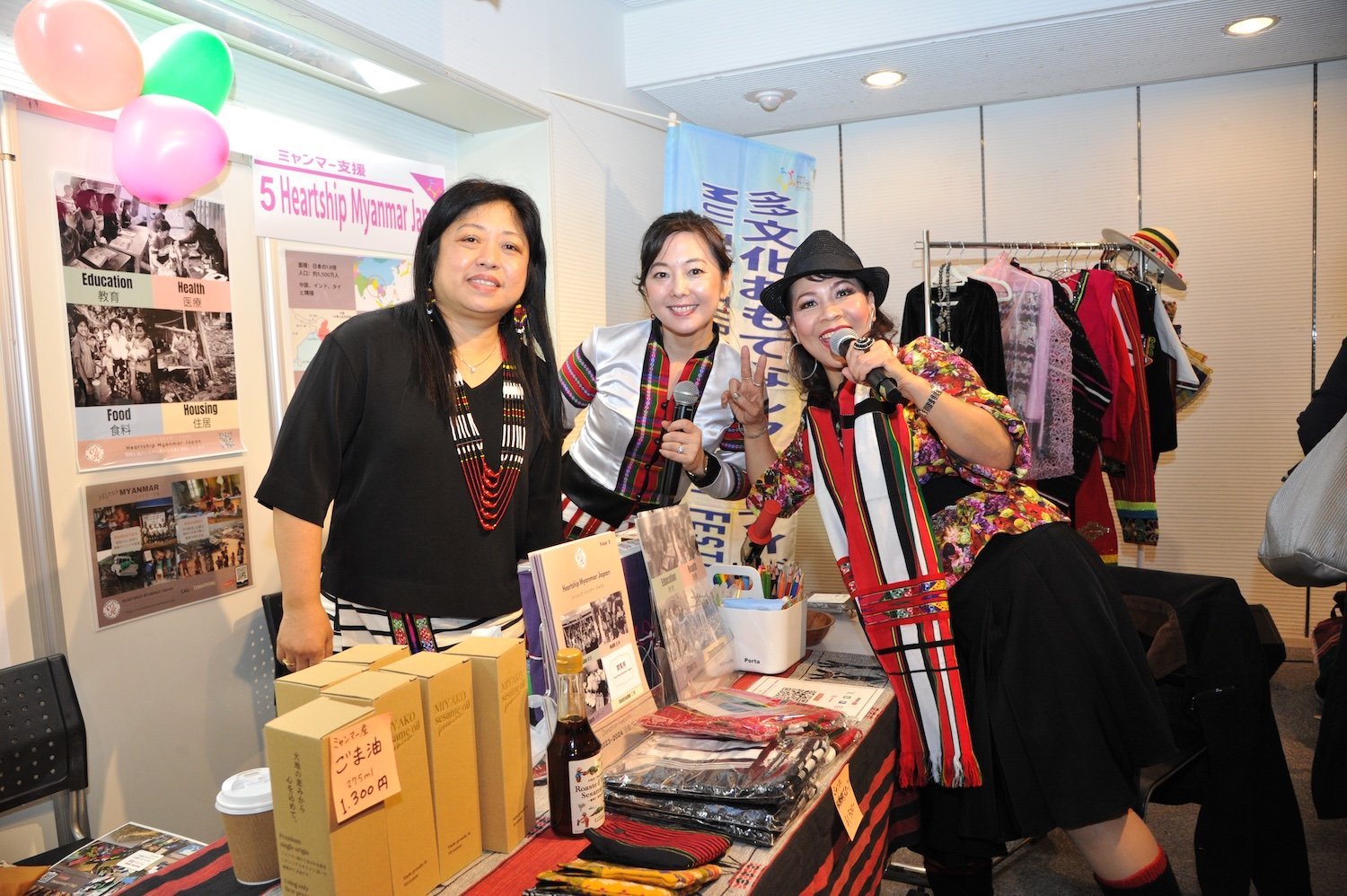 Heartship Myanmar Japan Three women in traditional clothing stand at a booth, their joyful spirits connecting smiles as they showcase cultural items and share insights. It’s a vibrant scene at this multicultural family festival, celebrating diversity and community.