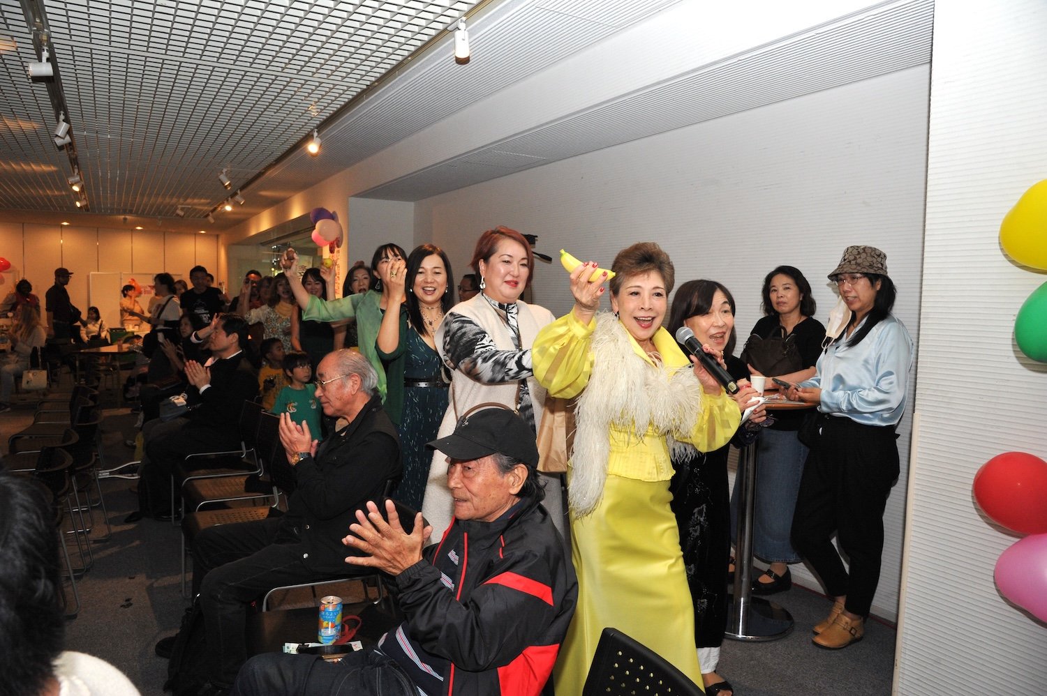 A multicultural group of people, some standing and some sitting, are gathered in a room with colorful balloons. At this lively 2024 family festival, a woman in a yellow dress smiles warmly and gestures toward the camera.