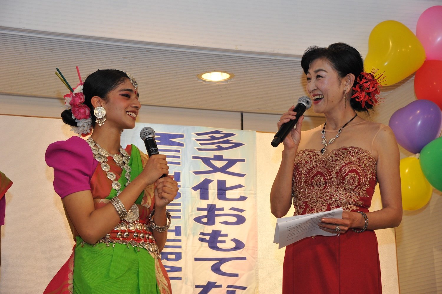 Two women on stage smile while speaking into microphones at a vibrant 2024 multicultural event. One wears traditional attire with vivid colors, the other dazzles in a red lace dress. Balloons float joyfully in the background, capturing the festive and inclusive spirit of this family festival.