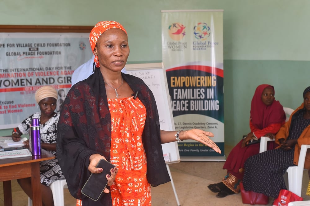 In a room adorned with banners on empowering families in peacebuilding, a woman addresses the audience passionately about uniting communities. Participants from Nigeria sit and listen attentively, eager to learn how they can combat gender-based violence together.