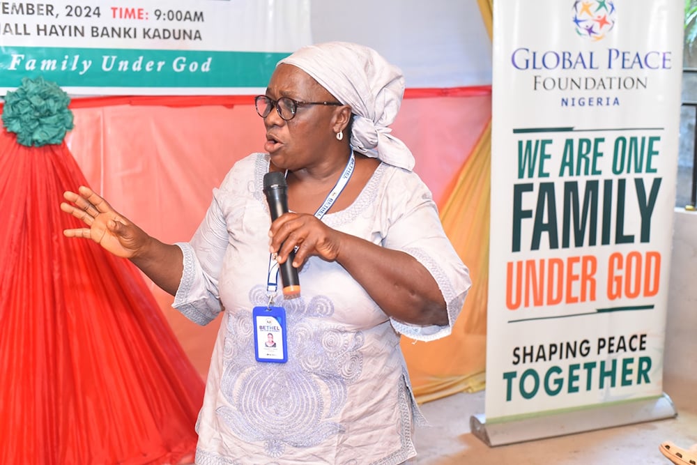 An elderly woman passionately speaks into a microphone at a Global Peace Foundation event, with a banner reading