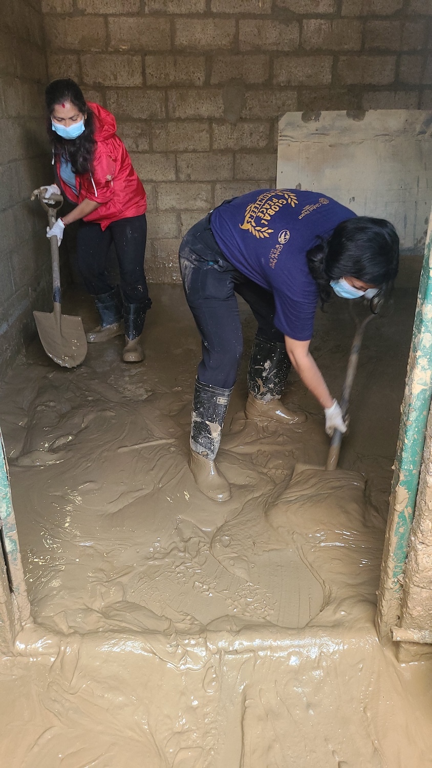 Two people wearing masks, gloves, and boots are shoveling mud inside a partially constructed room with brick walls, remnants of the recent Balkhu Flood that hit Kathmandu.