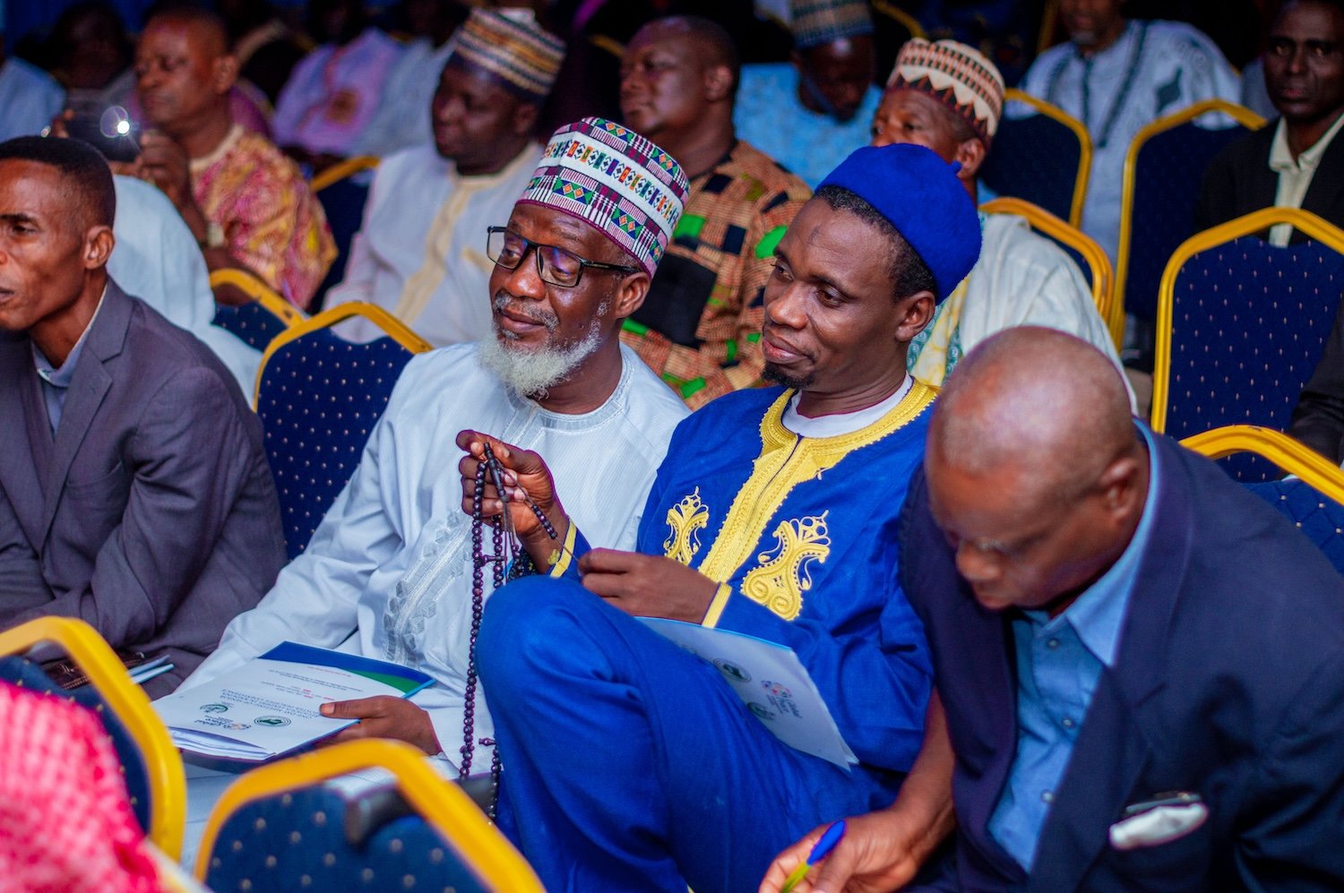 A group of men in traditional and formal attire sit in a row during the Kaduna Peace Conference, with some holding programs.