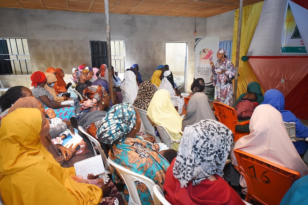 At a Hayin Banki Community workshop, a group of women in colorful headscarves gather in a room with banners and chairs. They focus on conflict analysis as a speaker leads the session, while one woman gently holds her baby, all united in their quest for sustainable peace.
