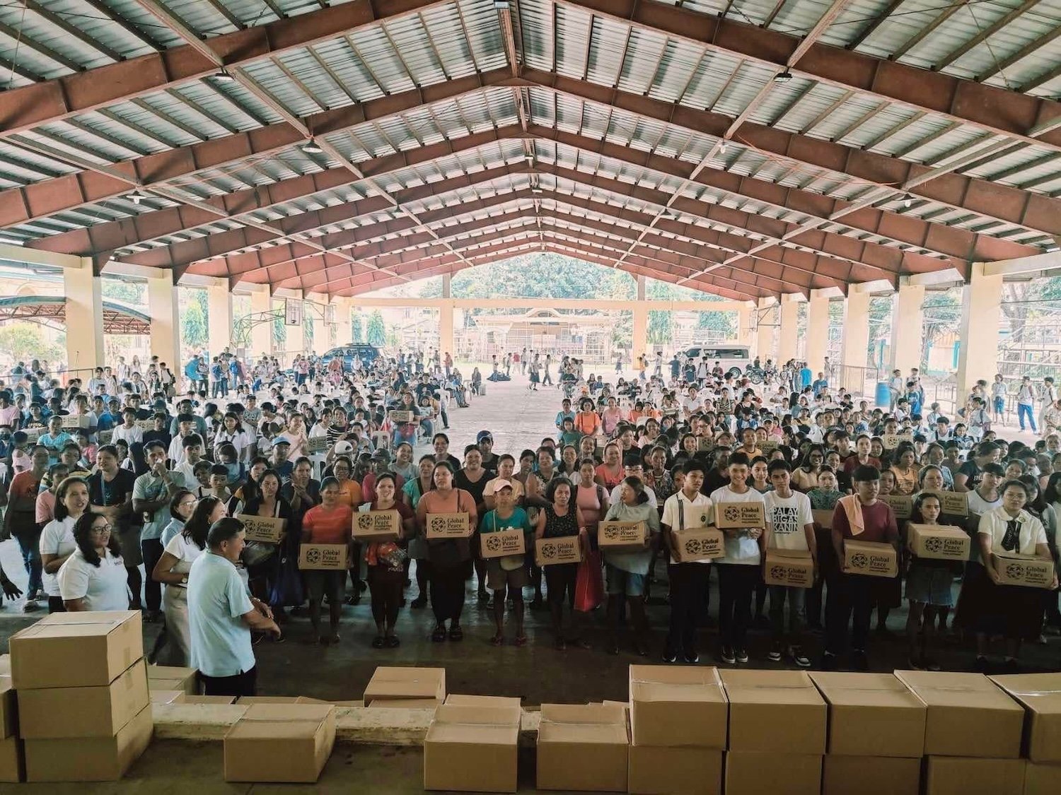 A large group of people stands in a covered area, holding boxes labeled