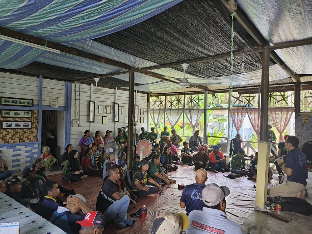 A group of Malaysian farmers gather indoors on a floor covered with mats, attentively listening to a speaker in a community setting about the benefits of syntropic agroforestry.