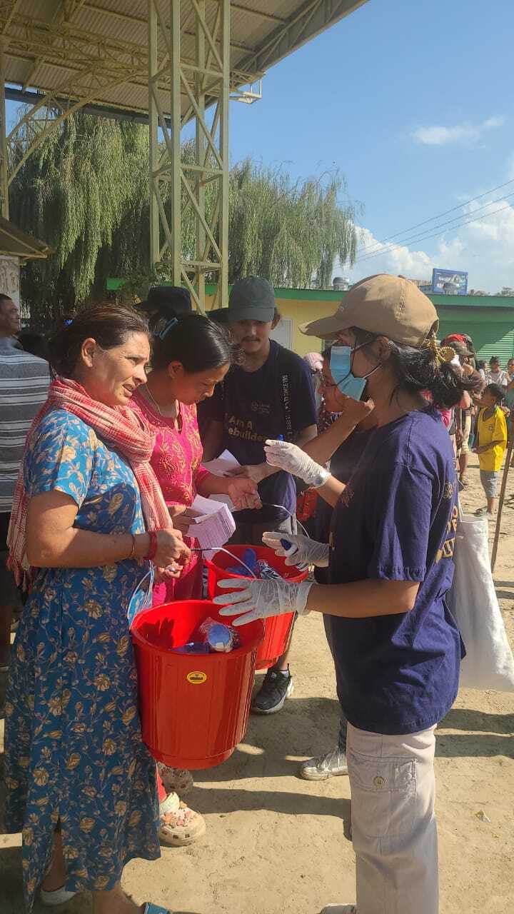 Amid the Balkhu Flood Crisis, people show resilience as they distribute red buckets to a group of women outdoors. Some individuals wear gloves and face masks as a safety measure, embodying the community spirit fostered by GPF Nepal.