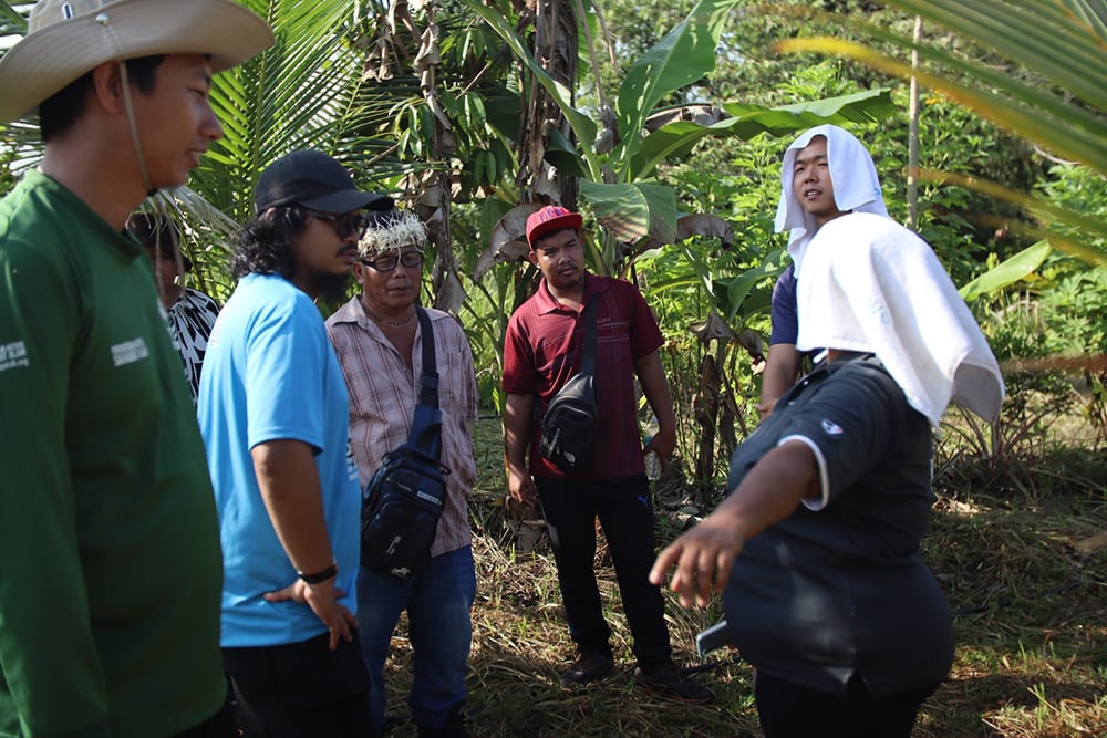 A group of six people, including Malaysian farmers, stands outdoors among tropical plants, engaged in conversation. One person uses a towel for shade. They are dressed in casual attire and hats, possibly discussing techniques like Syntropic Agroforestry in their GPF-led initiative.