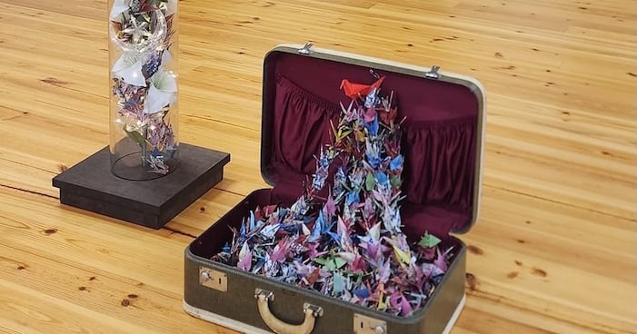 A suitcase overflowing with vibrant Montana Peace Cranes sits on the wooden floor, beside a cylindrical glass container filled with more cranes, symbols of hope and harmony inspired by the Cross Community Reconciliation Program.