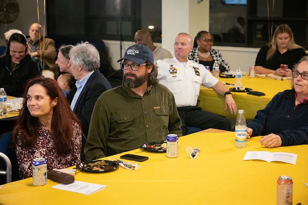 People sitting around tables covered with yellow tablecloths, eating and drinking, at an indoor event.