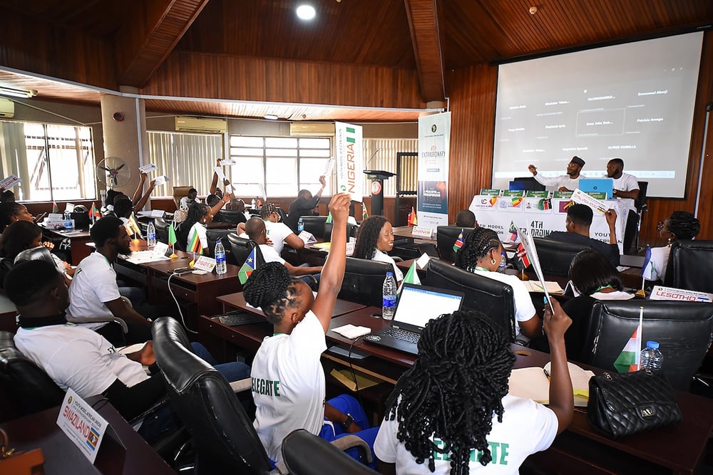 A classroom setting with students raising hands and engaging in discussion. Two presenters at the front have a projector displaying slides.
