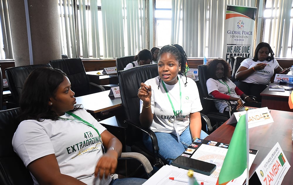A group of people in a conference room sit at desks, wearing matching shirts and badges. Two women in the foreground are engaged in conversation. A banner for a peace foundation is visible.