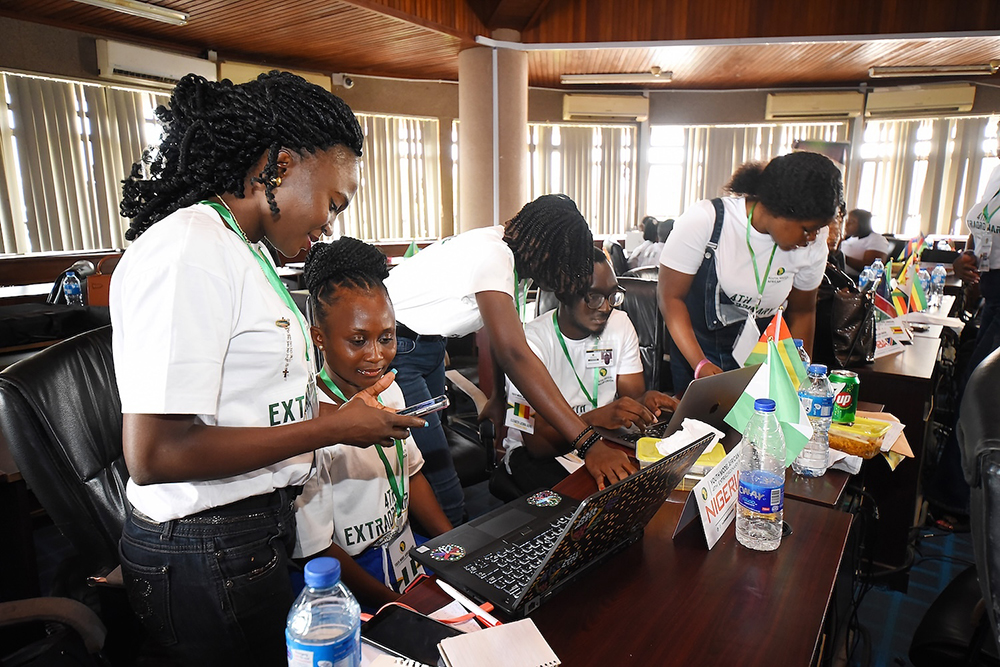 Group of people in white shirts working together on laptops at a conference table with flags and water bottles.