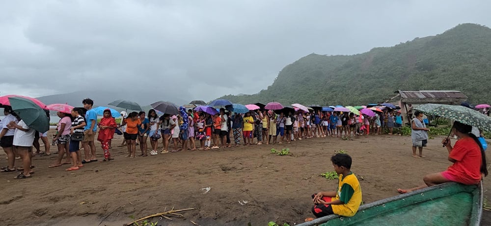 A large group of people stand under colorful umbrellas on a cloudy day near a mountain in Flooded Camarines Sur. Some are sitting on the ground, with a wooden structure in the background as they unite to Rebuild Communities.
