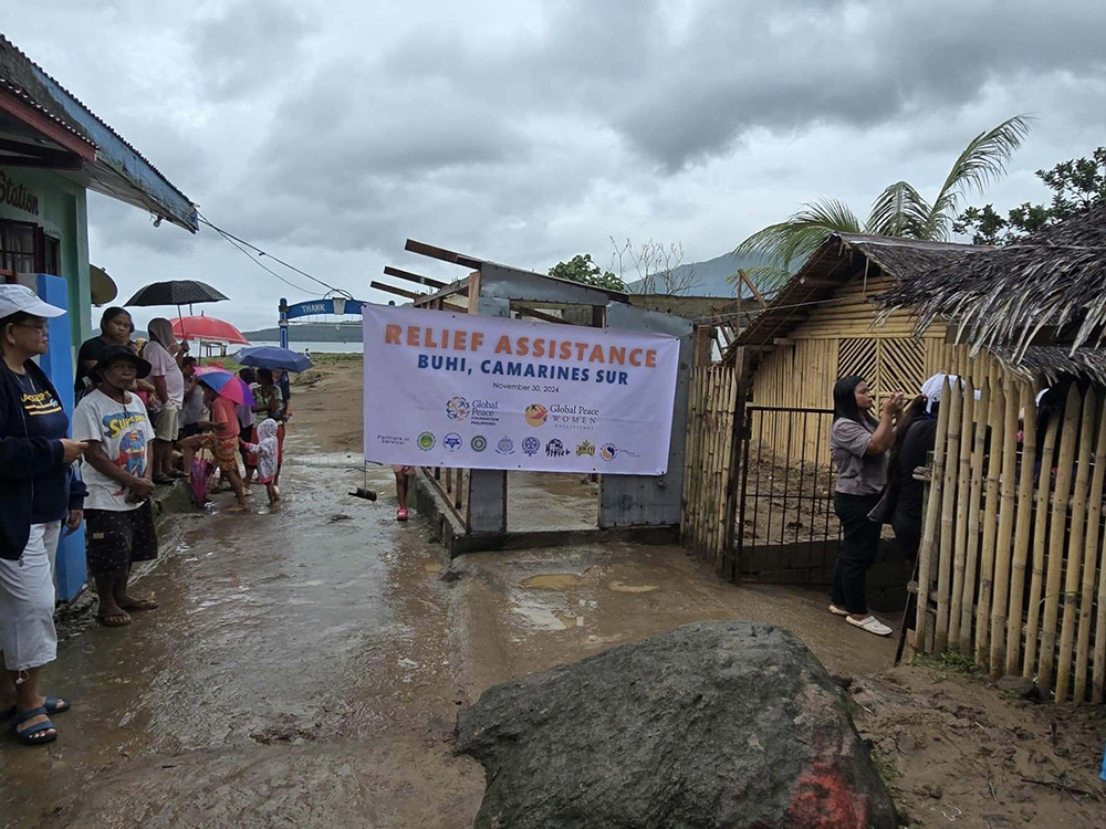 People standing in a muddy area near a banner reading "Relief Assistance Buhi, Camarines Sur" under a cloudy sky, as part of the GPF Philippines' efforts to rebuild the community.