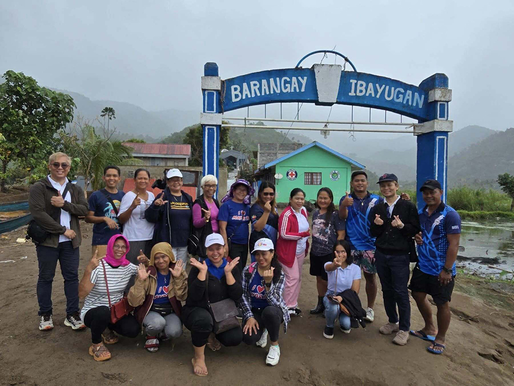 A group of people from Camarines Sur Communities poses in front of the Barangay Ibayugan entrance sign, proudly showcasing their efforts to rebuild. A small building and mountains are visible in the background, symbolizing the hope and promise led by GPF Philippines.