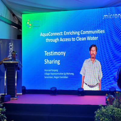 Untitled-design-2 A speaker on stage stands next to a large screen displaying a slide titled "Testimony Sharing" with the topic "AquaConnect: Enriching Communities through Access to Clean Water," highlighting how Micron and Global Peace partner to bring clean water to Orang Asli communities.