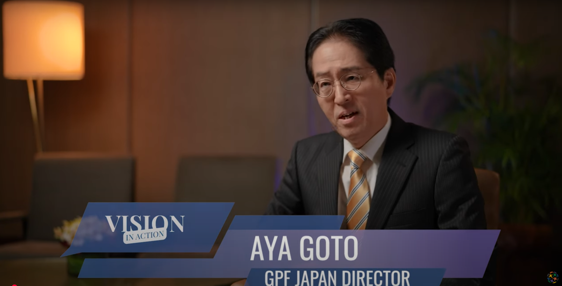 A person in a suit and glasses is speaking in an office setting, embodying the theme of "Vision in Action." The text reads: "Aya Goto, GPF Japan Director," highlighting the drive towards creating a vibrant multi-cultural society.