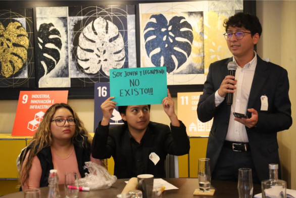 Three people sit at a table in a meeting room adorned with framed art and printed signage. One holds a sign saying, "Soy joven y el cambio no existe!!!" as they discuss initiatives with the Global Peace Foundation's Central America team.
