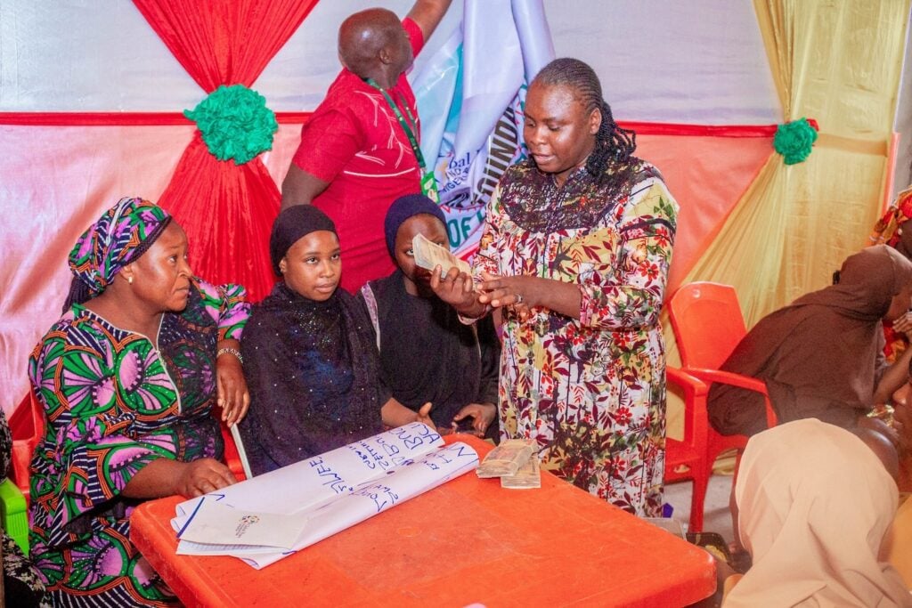 A woman in a colorful dress stands at an orange table with seated women, holding a stack of money and engaging with the group, embodying the spirit of women empowerment fostered by Cooperative Societies like GPF Nigeria.