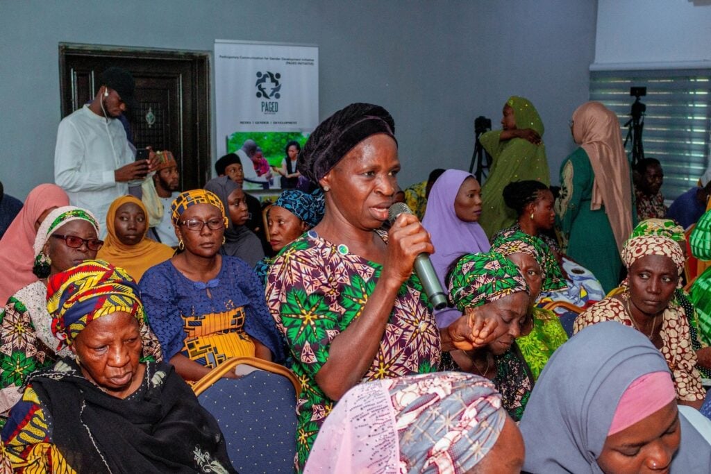 A woman speaks into a microphone while seated, surrounded by other women in colorful clothing at a conference setting. Celebrating International Women's Day, they unite to discuss how women lead the way towards lasting peace in Nigeria.