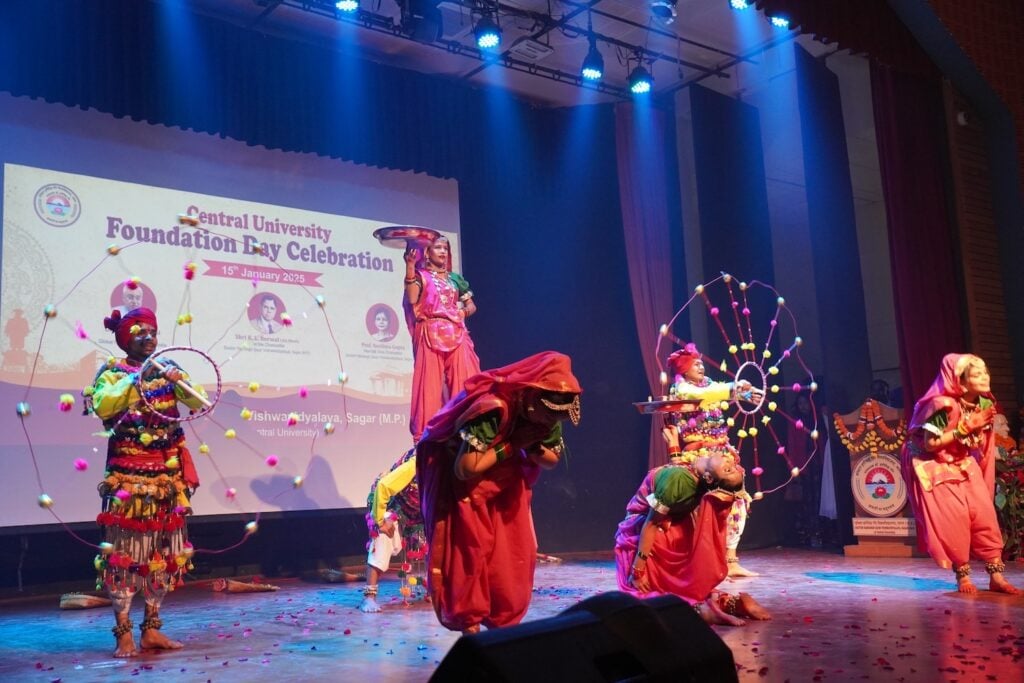 Performers in colorful traditional attire dance on stage at the Central University Foundation Day Celebration, their graceful movements embodying a mission of spreading peace, set against a backdrop displaying event details at Dr. Harisingh Gour Vishwavidyalaya.