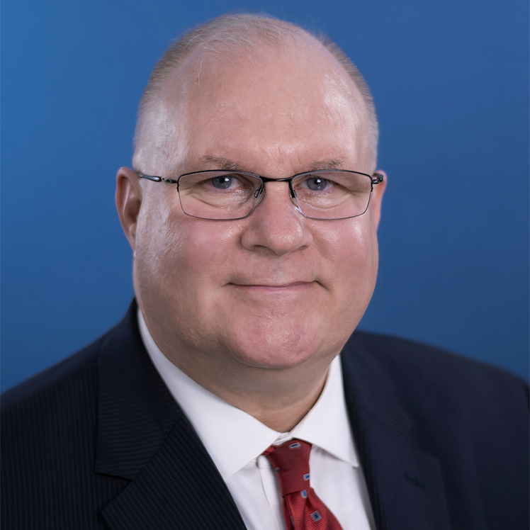 David Maxwell A man in a suit with a red tie is smiling against a blue background. He is wearing glasses and has short, light-colored hair.