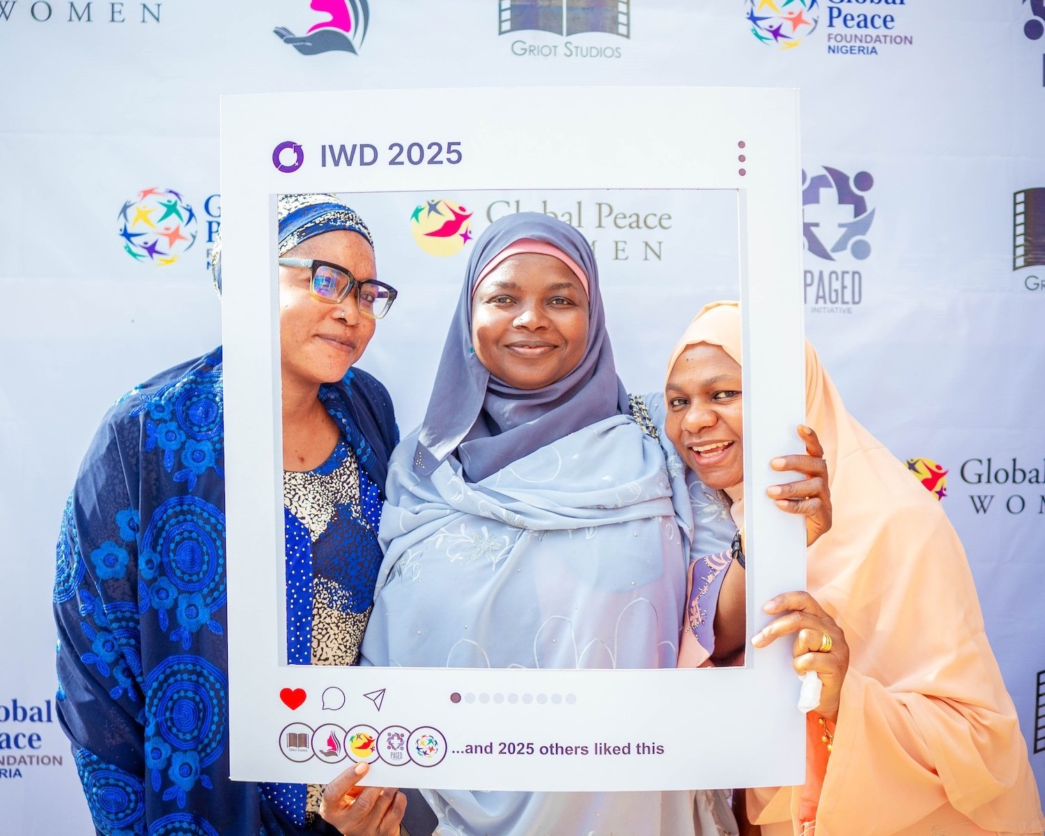 Three women in colorful hijabs pose together behind a large Instagram photo frame prop labeled "IWD 2025" at a Women’s Day event in Nigeria, with logos and banners promoting peace in the background.