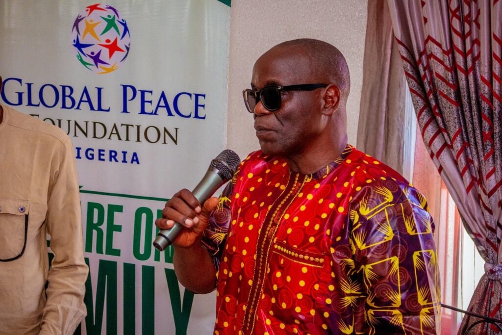 A man in patterned attire and sunglasses speaks into a microphone near a Global Peace Foundation Nigeria banner, highlighting peacebuilding initiatives.