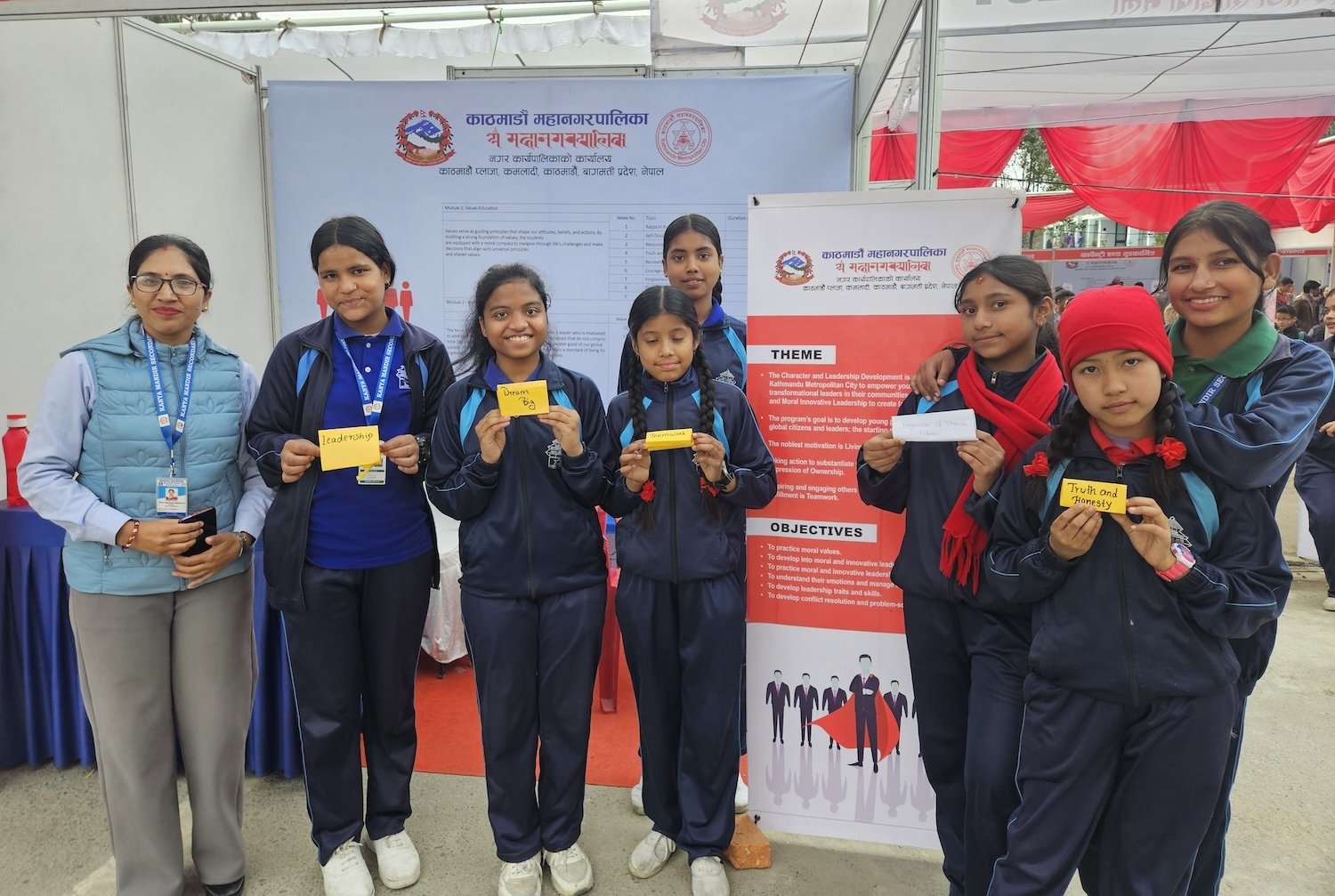 A group of people in navy tracksuits and a woman in a light blue jacket, representing the Department of Education, pose in front of a booth displaying educational materials and holding yellow or white paper slips at an event in Kathmandu.