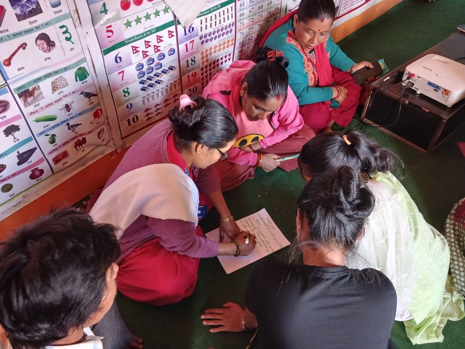 A group of women in Nepal sit on the floor, collaboratively writing on a sheet of paper, with educational posters displayed on the wall beside them—demonstrating women empowerment and peaceful dispute resolution.