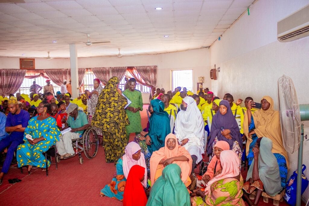 A group of people, including women in colorful hijabs, men in patterned clothing, and people with disabilities, gather inside a bright, spacious hall with curtains and ceiling fans for a peacebuilding event.