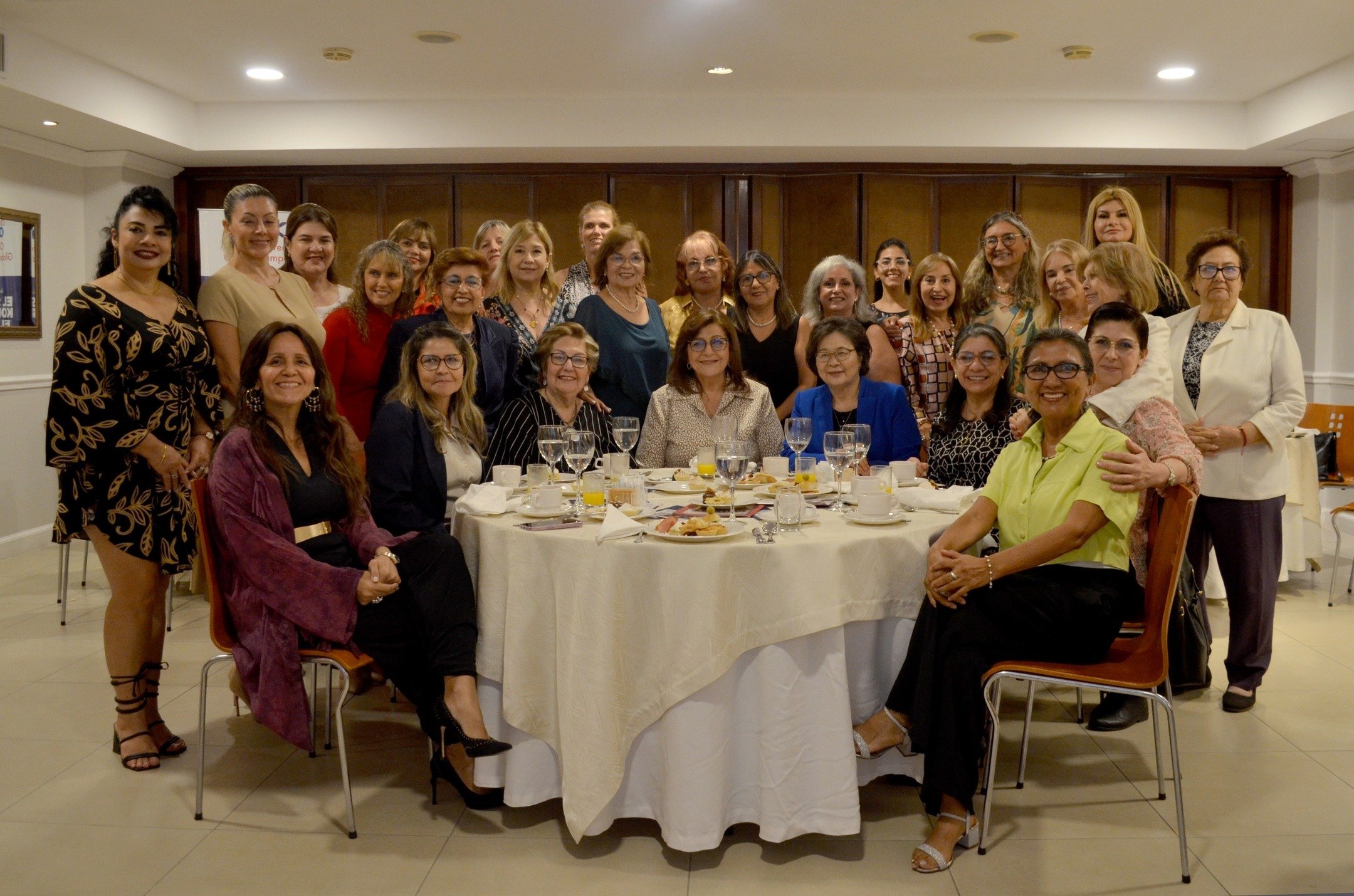 A group of women pose for a photo around a round dining table set with glasses, plates, and food in a well-lit indoor setting, celebrating International Women’s Day with Area Mujer Paraguay.