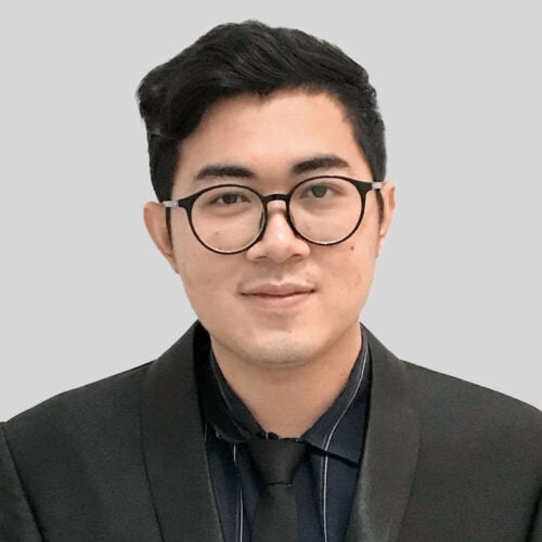A man with short black hair and glasses, wearing a dark suit, black shirt, and tie, poses in front of a plain light gray background.