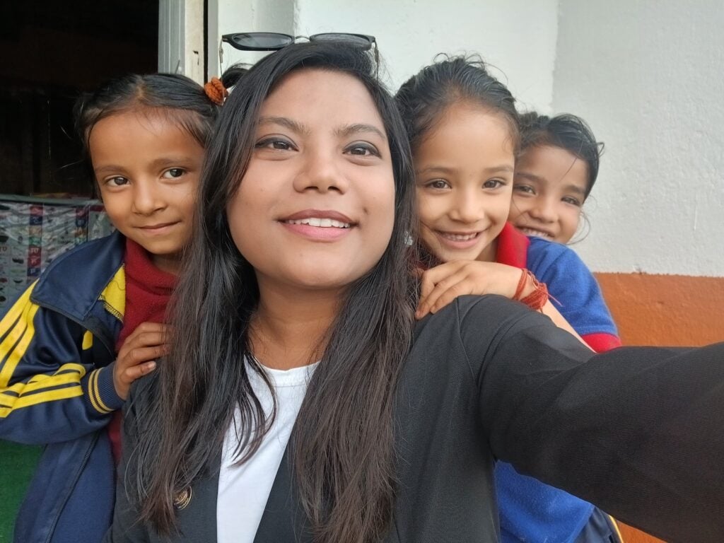 A woman, embodying women empowerment, takes a selfie with three smiling children standing close behind her in front of a white and orange wall in Nepal.