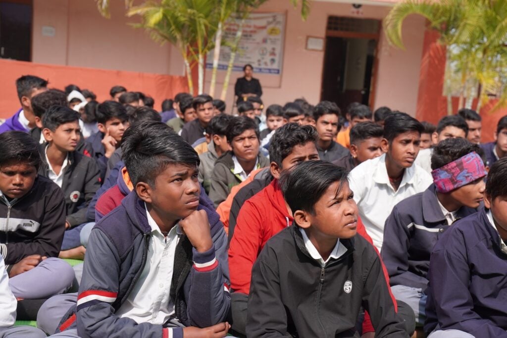 A group of students sits attentively outdoors, proudly wearing their school uniforms. In the background, a poster displayed on a building wall subtly conveys a message of peace and the mission of Dr. Harisingh Gour Vishwavidyalaya.