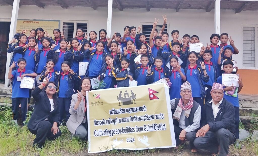 A large group of students and adults pose together holding a banner that reads “Cultivating peace-builders from Gulmi District, 2024” in front of a building in Nepal. Some hold certificates, celebrating peaceful dispute resolution and women empowerment.