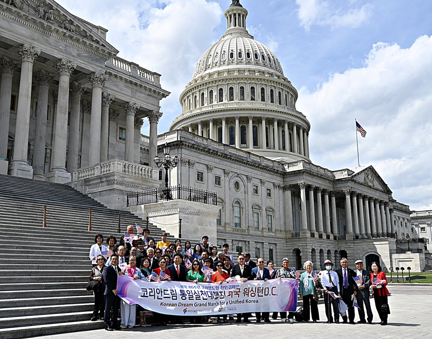 A large group of people stand on the steps of the U.S. Capitol during the Capitol Forum, holding a banner that reads "Korean Dream Grand March for Unified Korea" in Korean and English.