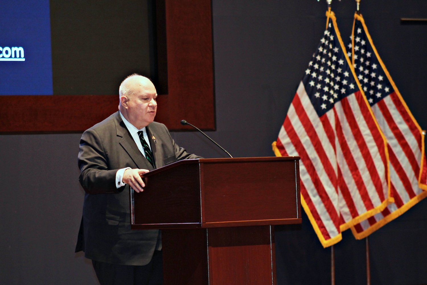 A man in a suit stands at a podium giving a speech at the Capitol Forum, with two American flags and a large screen in the background.