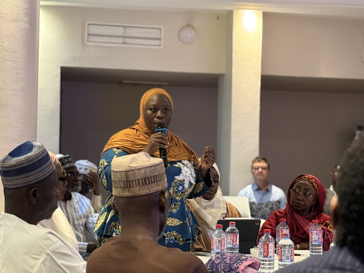 A woman in a hijab speaks into a microphone while standing at a table surrounded by seated people, water bottles, and papers in a West Africa conference room focused on Freedom of Religion and Belief.