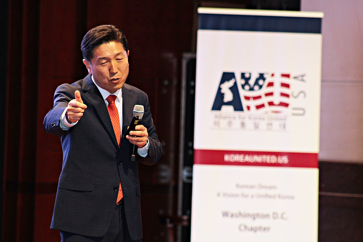 A man in a suit speaks and gestures while standing next to a sign for the Alliance for Korea United USA at a Capitol Forum event.