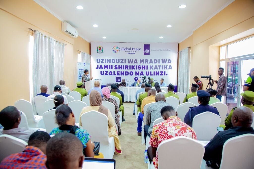 A group of people sit facing a panel at a community project launch event in Southern Tanzania, with banners and speakers at the front of the room, highlighting the importance of community collaboration.