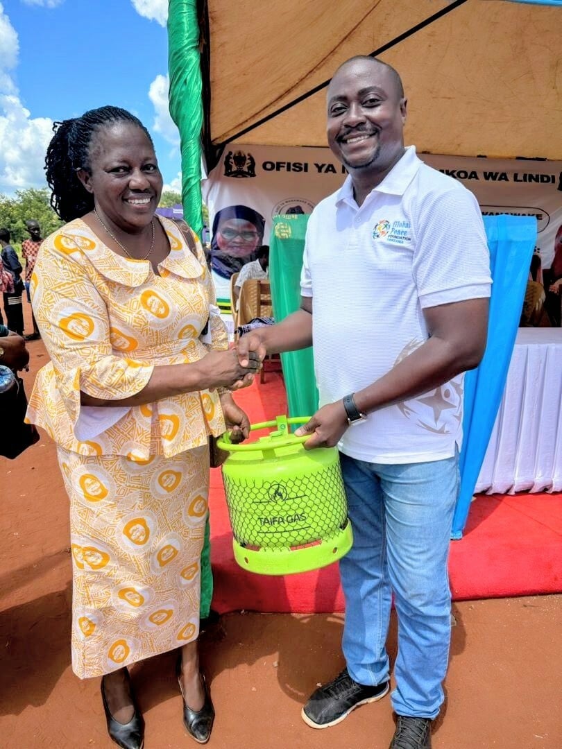 A woman and a man shake hands while holding a small green gas cylinder at an outdoor event with a tent and banners in the background.