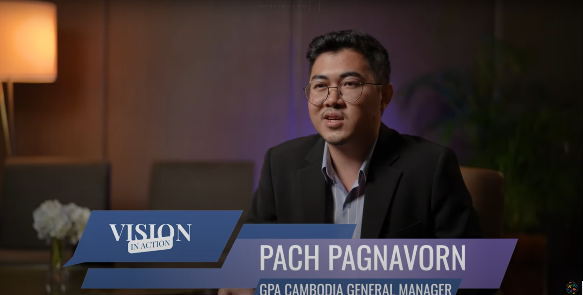 A man wearing glasses and a black suit sits indoors, speaking to the camera about Vision in Action. On-screen text reads: "VISION IN ACTION" and "PACH PAGNAVORN, GPA Cambodia General Manager," inspiring Cambodian youth toward peace.