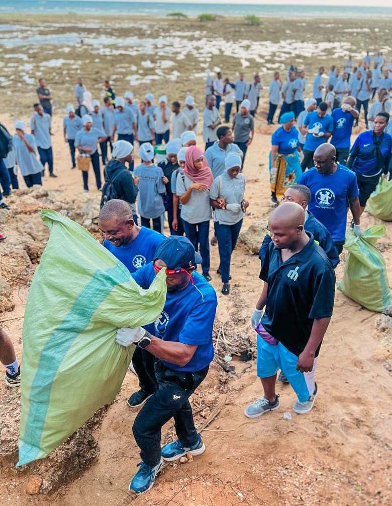 Demonstrating community power, a large group of people in Tanzania participate in a beach cleanup, collecting litter in large green bags near the shoreline to combat coastal pollution.