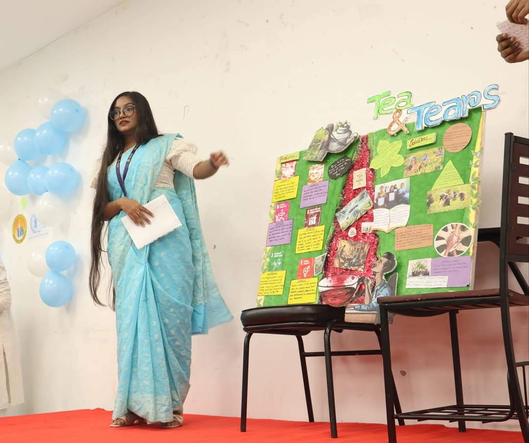 A woman in a blue sari stands on stage beside a colorful project board titled "Tea & Tears," showcasing youth leadership in Bangladesh, with balloons and a white wall in the background.