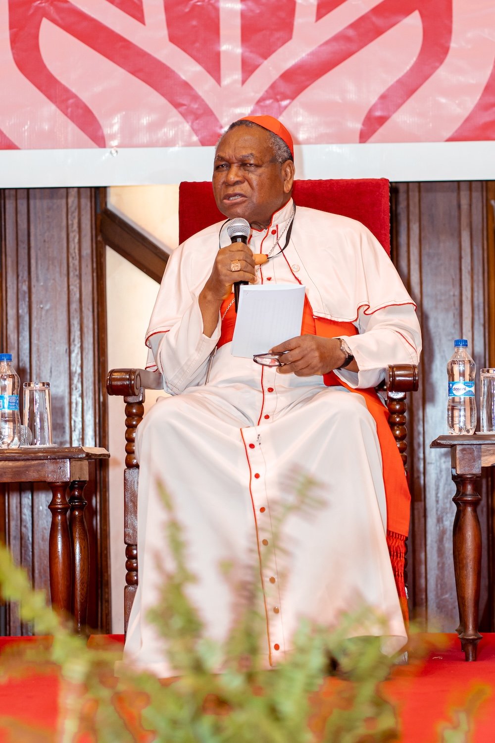 A man in clerical attire sits on a chair, holding a microphone and notepad, with water bottles nearby and a decorative backdrop behind—an engaging scene from the Global Peace Foundation’s IRF Summit Africa on religious freedom.