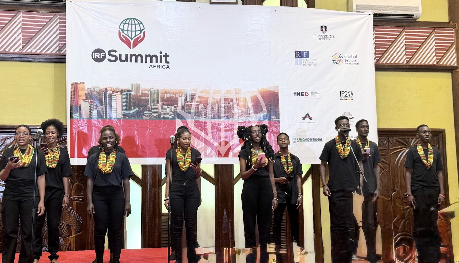 A group of performers in matching black outfits and colorful scarves sing on stage at the IRF Summit Africa, an event dedicated to religious freedom, with a large event banner behind them.