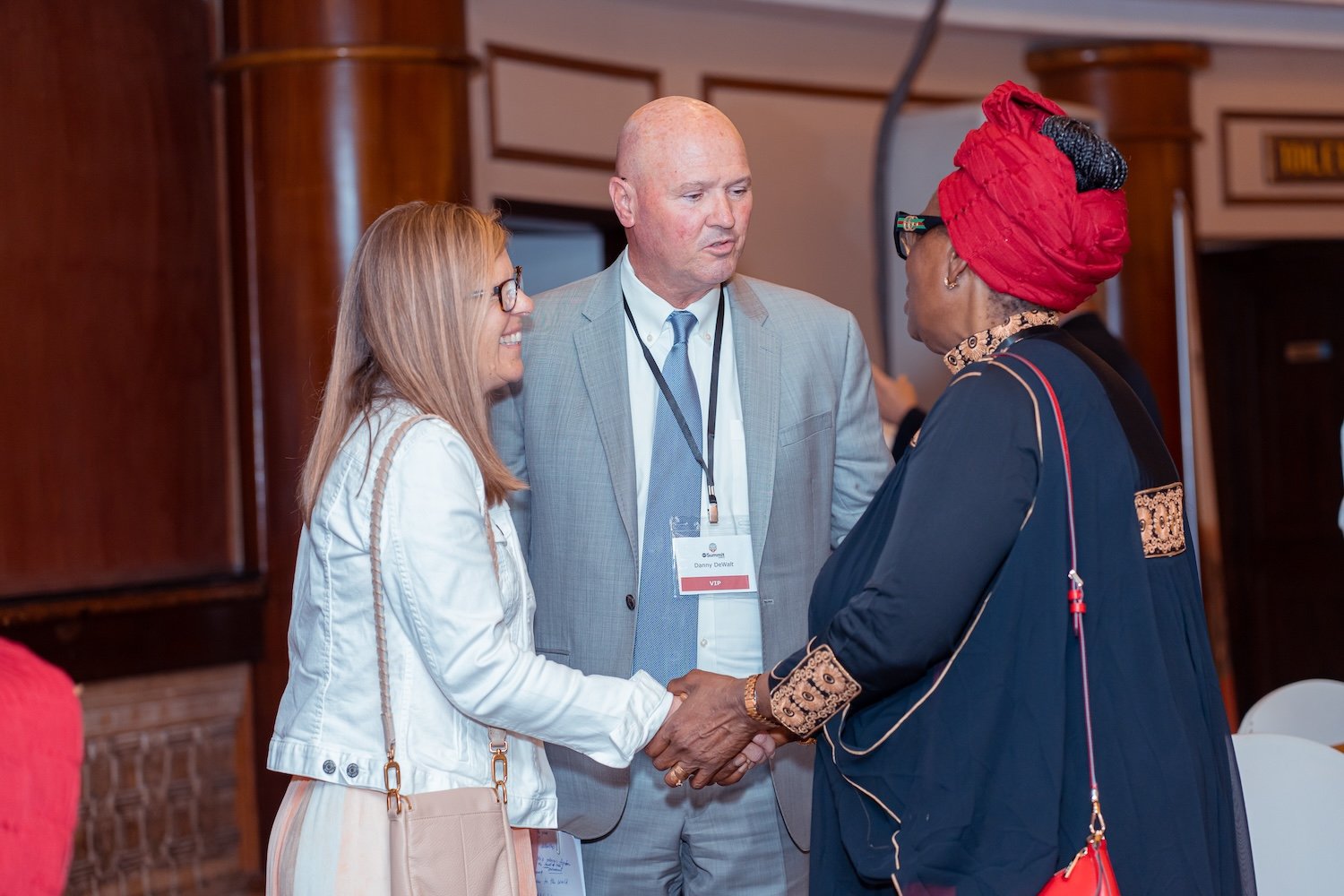 Three people in business attire stand indoors, two women and one man, engaged in conversation and shaking hands at a professional event focused on International Religious Freedom.